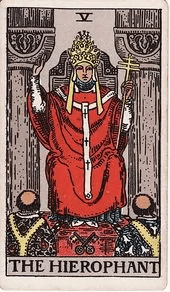 The Hierophant sits between two pillars in a triple-tiered crown, one hand raised in blessing over two kneeling acolytes.