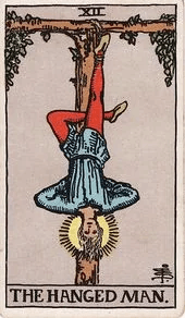A man hangs upside down by one foot from a wooden beam, his expression calm and still rather than distressed.